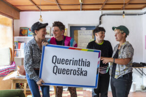 Kat, Mika, Lux und Lie halten ein Schild mit der Aufschrift Queerinthia/Queeroška.