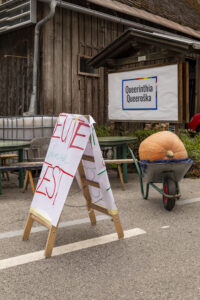 Festival-Schild an der Straße, im Hintergrund ein riesiger Kürbis in einer Scheibtruhe und das Ortsschild Queerinthia/Queeroška.